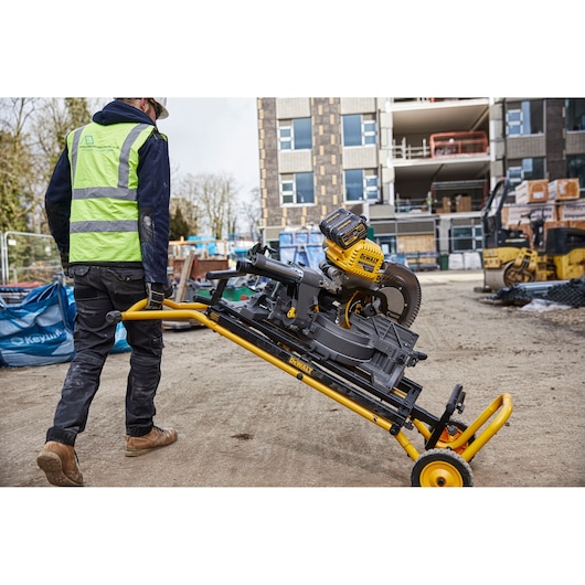 Worker moves a DEWALT miter saw on a rolling stand at a construction site with buildings in the background.
