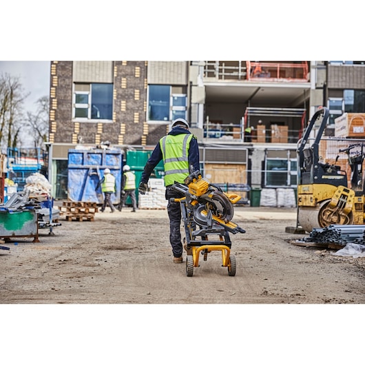 Worker moves a DEWALT saw on a wheeled stand at a busy construction site with other workers and building materials.