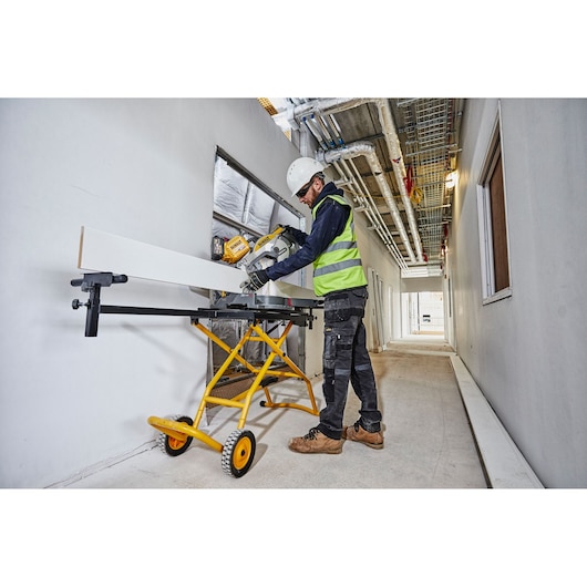 Person sawing wood on a DEWALT yellow mobile stand in a construction hallway, wearing safety gear.
