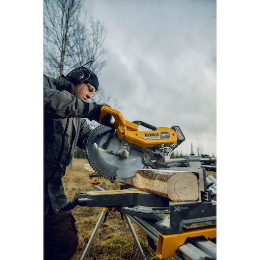 A DEWALT DHS780N saw cutting wood outdoors. Operator is wearing protective gear. Tools and log are visible.