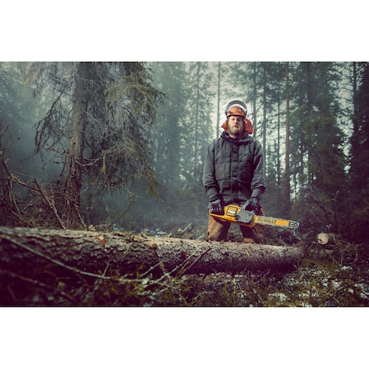 Person with blurred face holds a DEWALT chainsaw in a forest, next to a fallen tree, wearing safety gear.