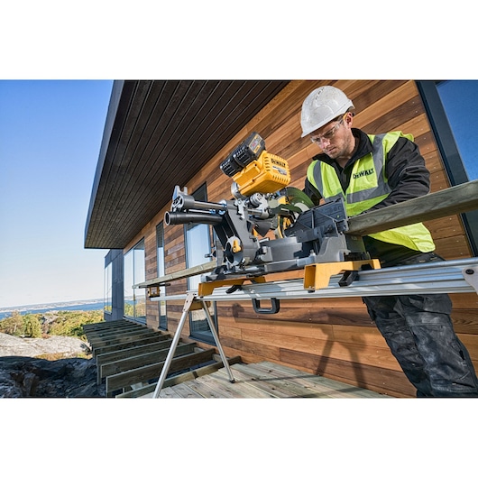 DEWALT miter saw in use outdoors, worker in safety gear cutting wood near modern house.