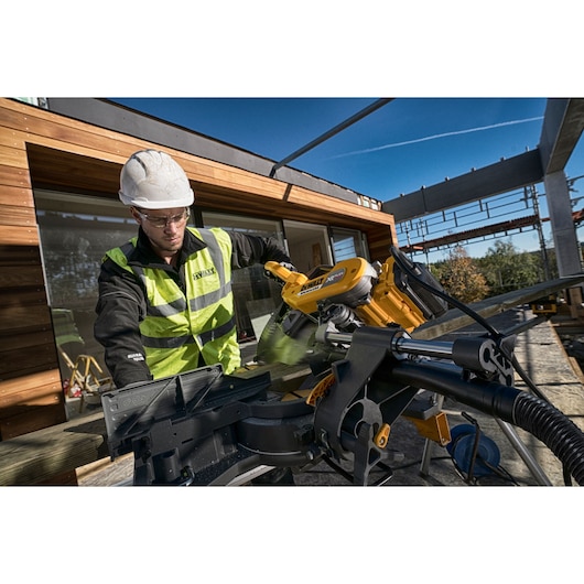 Worker uses a DEWALT DHS780N saw to cut wood on a construction site outdoors, wearing safety gear.