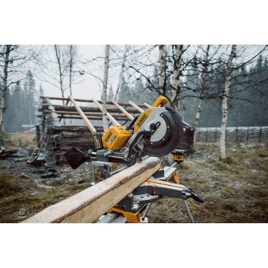 DEWALT miter saw cutting a wooden beam outdoors, with a rustic cabin and trees in the background.
