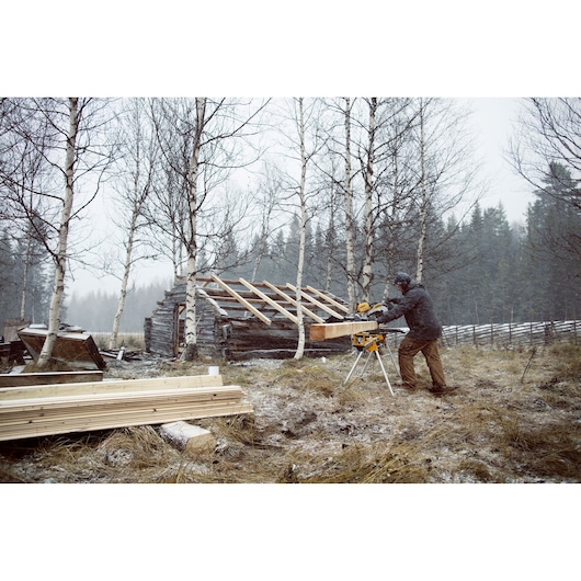 Person using a DEWALT saw outdoors to cut wood, near a cabin under construction in a snowy forest landscape.