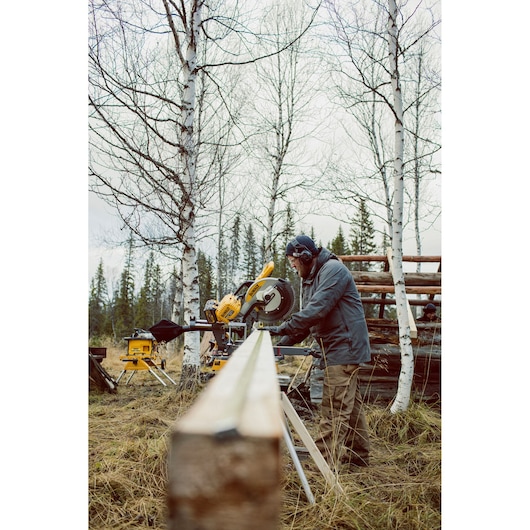 Person cutting wood outdoors with a DEWALT saw among birch trees; cold weather clothing and safety gear worn.