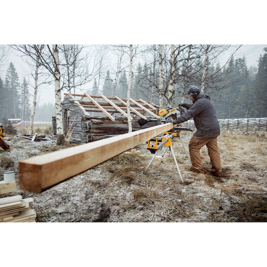 Man using DEWALT miter saw outdoors to cut long wooden beam near a cabin with snowy ground and trees.