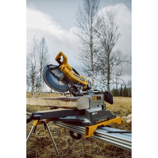 DEWALT cordless mitre saw cutting wood outdoors in a field with leafless trees and cloudy sky.