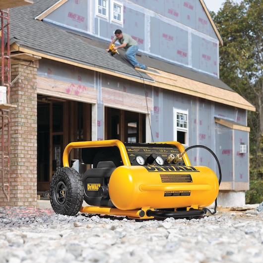 A DEWALT DPC17PS portable air compressor positioned on gravel at a construction site, with a person using a power tool on the roof of a partially built house.