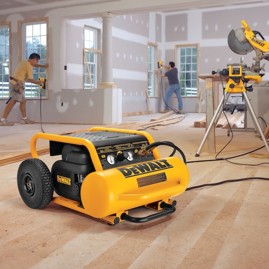A DEWALT portable air compressor (model DPC17PS) is positioned on the floor of a construction site, with workers installing window frames and other DEWALT tools visible in the background.