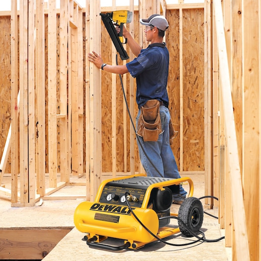 A construction worker uses a DEWALT air compressor and nail gun to build a wooden frame structure. The worker is standing among unfinished wooden walls, wearing a tool belt and cap. The yellow DEWALT compressor is placed on the ground with cables attached to the nail gun.