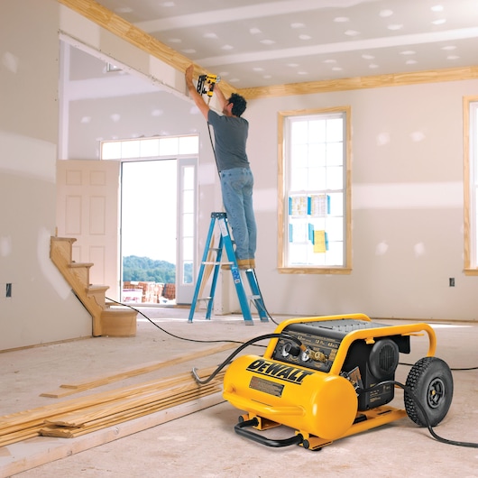 A portable DEWALT air compressor is placed on the floor of a room under construction, with a person standing on a ladder using a power tool to install wooden trim near the ceiling.