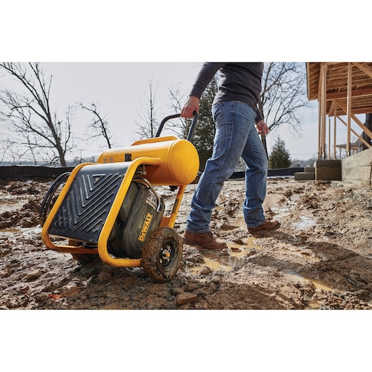 A portable DEWALT air compressor on wheels being pulled by a person across a muddy construction site. The person is wearing jeans and work boots.