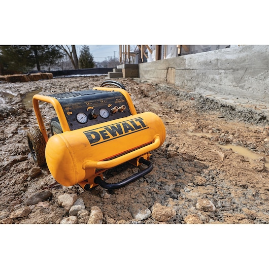 A DEWALT portable air compressor model DPC17PS in yellow and black, positioned on a muddy construction site with building materials and unfinished concrete structures visible in the background.