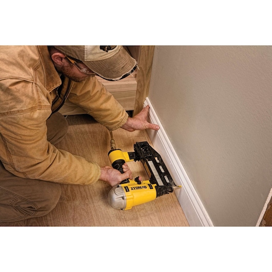 Person uses DEWALT nailer to attach trim to wall in a room with wood flooring.