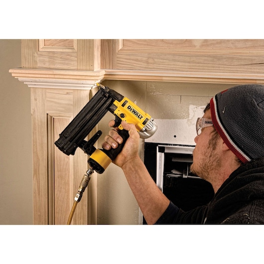 Person using a DEWALT nailer to attach wood trim to a wall, wearing safety glasses and a beanie.