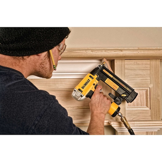 Person using a DEWALT nail gun to attach trim to wood paneling indoors.