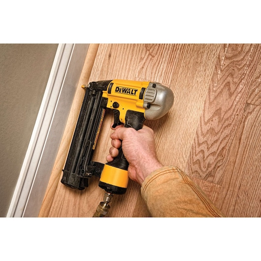 Hand using a DEWALT nailer tool on a wooden floor next to a wall.