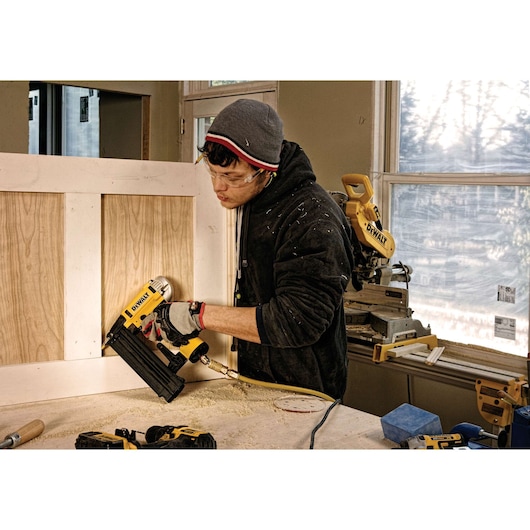 Person using DEWALT nailer to attach panel in workshop. Other DEWALT tools are visible on the table and background.