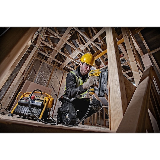 Worker uses DEWALT nail gun with compressor in a wooden construction site, wearing safety gear.