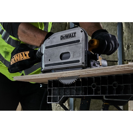 DEWALT circular saw is cutting plywood on a workbench, operated by a worker with gloves and safety vest.