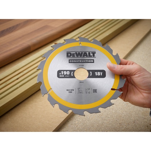 Hand holding a DEWALT DT1943 circular saw blade, 190 mm, 18 teeth, with wood boards in the background.