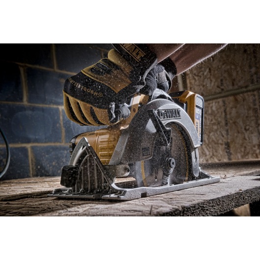 Gloved hand using a DEWALT circular saw to cut wood, with sawdust flying in a workshop setting.