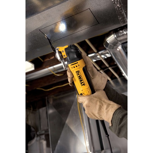 Gloved hands use a DEWALT oscillating tool to cut a rectangular opening in a metal duct in an industrial setting.