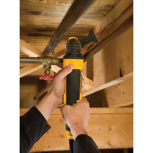 Hands using a DEWALT power tool to cut a metal pipe under a wooden ceiling structure.