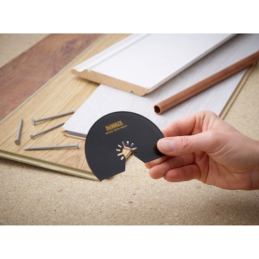 Hand holding DEWALT DT20708 saw blade, with wood, nails, and pipe in background.
