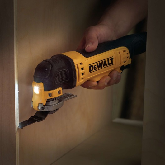 A DEWALT multitool is used to cut wood, with a hand holding it and LED light illuminating the cutting area.
