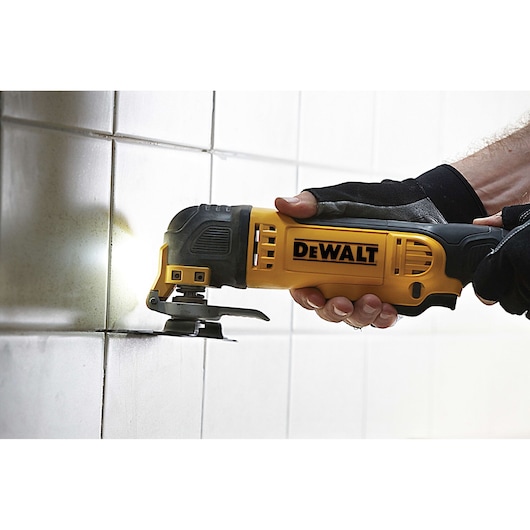 DEWALT oscillating multi-tool cutting tile grout, held by gloved hand against white tiled wall.