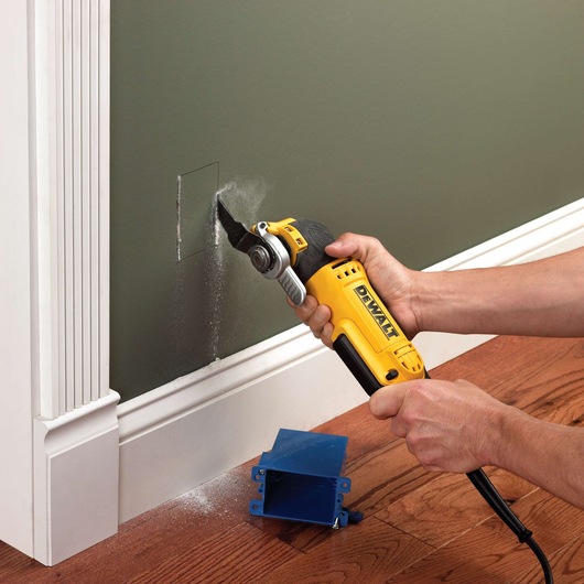 DEWALT tool cutting a hole in a wall near the floor, with a blue electrical box and sawdust visible.