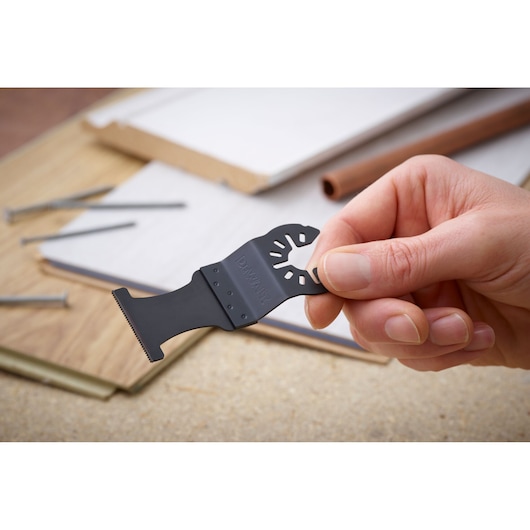 Hand holding a DEWALT DT20743 oscillating multi-tool blade above work materials and nails.