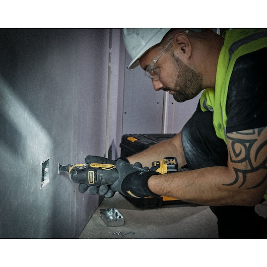 Person using DEWALT multi-tool to cut drywall. Tool case and screws are visible on the floor nearby.