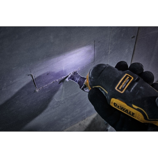 DEWALT tool cutting a rectangular hole in drywall, held by a gloved hand, with LED light illuminating the area.
