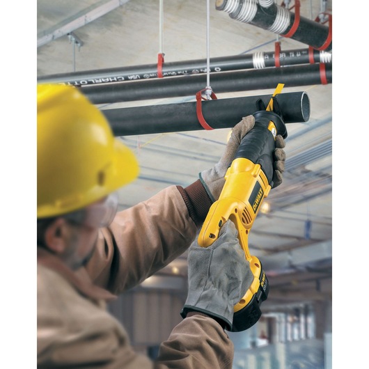 Worker uses a DEWALT reciprocating saw to cut a black pipe overhead in an industrial setting.