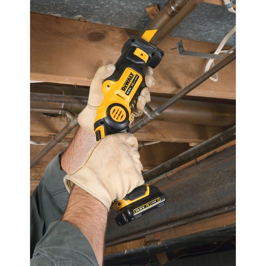 Person using a DEWALT cordless saw to cut a pipe under a wooden ceiling; wearing work gloves for safety.
