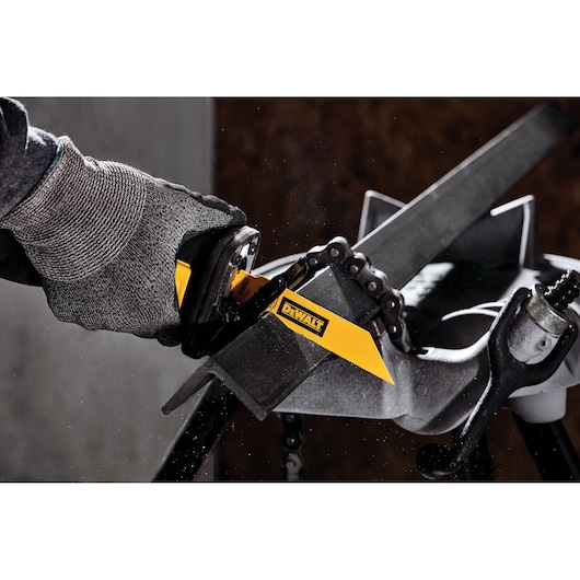 A person wearing safety gloves uses a DEWALT reciprocating saw blade to cut through a metal bar. The DEWALT blade is yellow with the brand logo clearly visible. The scene takes place in a workshop setting.