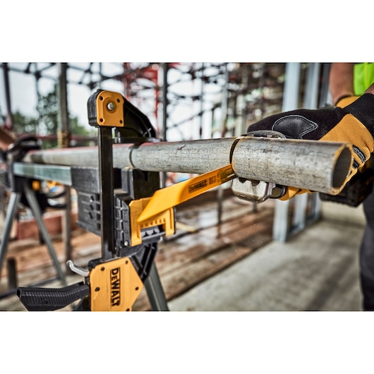 A DEWALT clamp securely holds a metal pipe on a workbench while a person wearing protective gloves uses a pipe cutter to cut through the pipe. The setting appears to be a construction site.