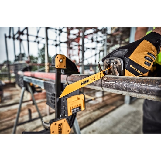 A DEWALT DT2416 reciprocating saw blade is cutting through a metal pipe on a construction site. The pipe is secured with clamps and a person wearing work gloves is operating the saw.