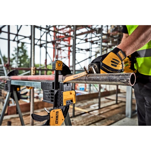 A worker wearing DEWALT branded gloves is using a DEWALT hacksaw to cut a metal pipe, which is secured in place with a DEWALT clamp, at a construction site with scaffolding in the background.