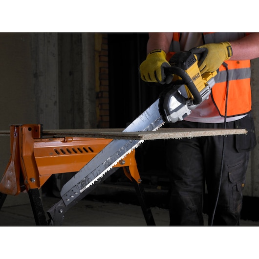 Person cuts wood with a DEWALT electric saw, wearing gloves and safety vest, wood supported by an orange workbench.