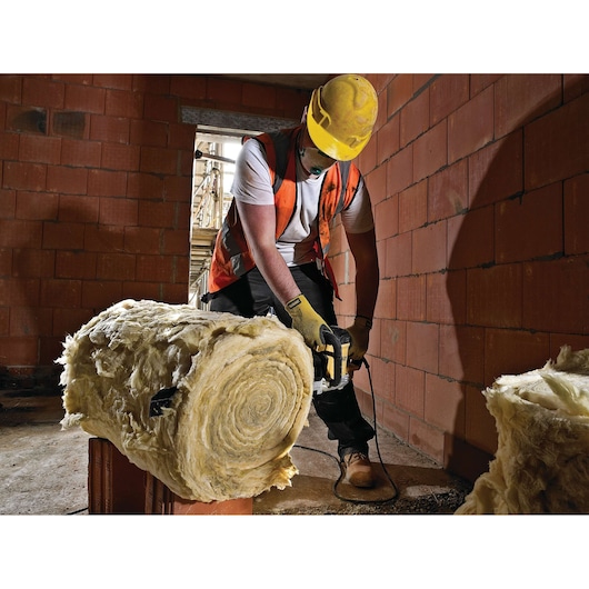 Worker uses DEWALT tool to cut insulation material in a construction site with red brick walls.