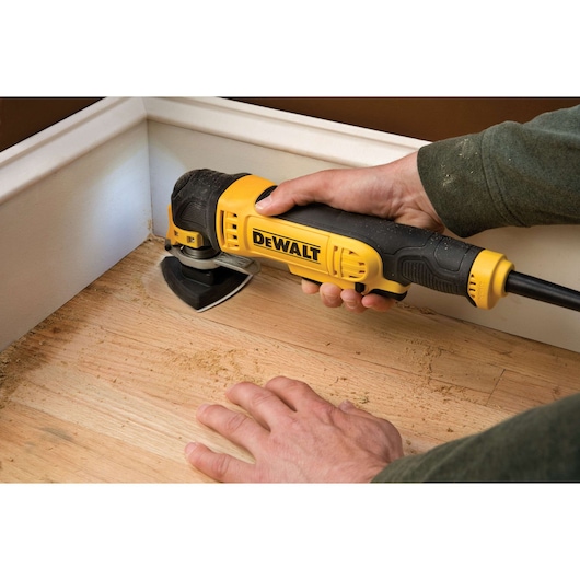 DEWALT sander being used on a wooden floor near a wall corner, held by a person's hand.