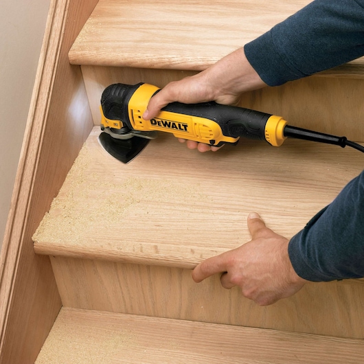 A DEWALT tool is used to sand wooden stairs, creating sawdust. Hands hold and operate the sander on a stair step.