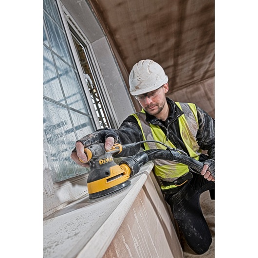 Worker uses a DEWALT sander to smooth a window ledge in a construction area, wearing safety gear and a helmet.