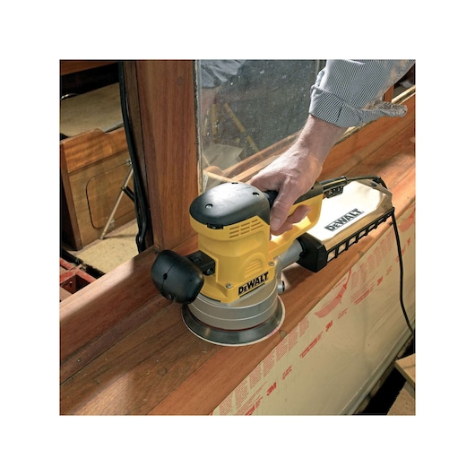 Person uses a DEWALT DT3123 sander to smooth wooden surface near a window. Tool is powered and yellow-black.