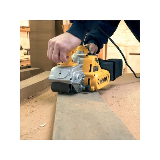 DEWALT belt sander in use, sanding a wooden plank in a workshop with sawdust visible on the surface.