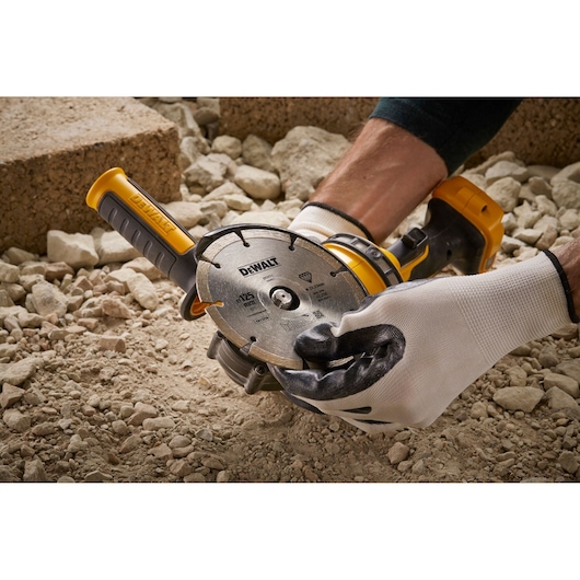 Gloved hands hold a DEWALT DT3559 cutting disc tool on rocky ground, showing its blade and yellow handle.